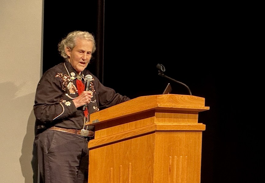 Temple Grandin stands at a wooden podium and speaks into a microphone.
